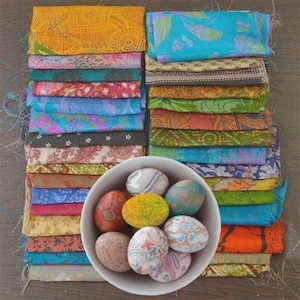 Puede incluir: Una colección de retazos de tela coloridos en varios patrones y colores, apilados ordenadamente. Un cuenco blanco contiene varios huevos de Pascua decorados. Las telas incluyen tonos de amarillo, azul, verde y naranja, creando una exhibición vibrante.