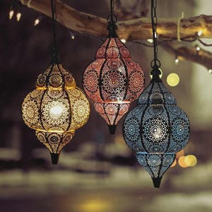 Op de afbeelding: Drie hangende decoratieve lantaarns met ingewikkelde, opengewerkte patronen. De lantaarns zijn in goud, rood en blauw, elk opgehangen aan een ketting. De achtergrond is wazig, met zachte verlichting en bokeh-effecten.