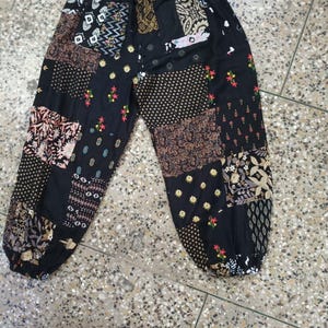 Op de afbeelding: Zwarte patchworkbroek met een trekkoord in de taille. De broek heeft verschillende stoffen vierkanten met patronen in zwart, bruin en crème. Het ontwerp bevat bloemen- en geometrische patronen, met rode en gele bloemenborduursels.