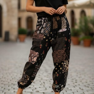 May include: Black patchwork trousers featuring diverse patterns, such as floral and geometric motifs. The trousers have a drawstring waist and are worn with a black t-shirt and beige slip-on shoes. The trousers are a casual style.