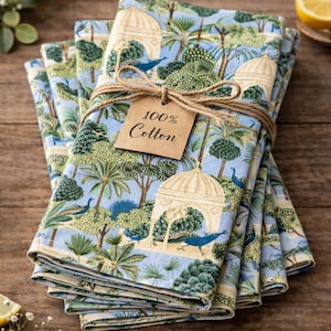 May include: A stack of folded cotton napkins tied with twine. The napkins feature a blue background with a repeating pattern of trees, gazebos, and peacocks. A tag reads "100% Cotton".