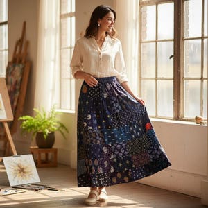 Puede incluir: Una mujer con una blusa crema y una falda larga azul marino con diseño patchwork. La falda tiene patrones florales y geométricos. Está de pie cerca de una ventana en una habitación luminosa. La falda mide unos 90 cm de largo.