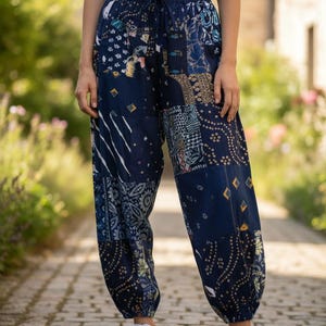 以下が含まれることがあります： ネイビーブルーのパッチワークパンツ。ウエストはドローストリング、足首はゴム入り。青、白、金色の様々な柄の生地パネルが特徴です。モデルは白いスニーカーを履いています。