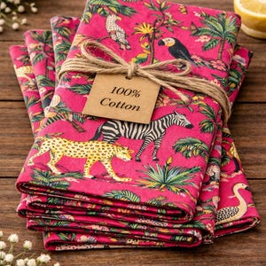 Op de afbeelding: Een stapel van vier roze katoenen servetten met een jungleprint met zebra's, luipaarden en vogels. De servetten zijn samengebonden met touw en een label leest "100% Cotton".