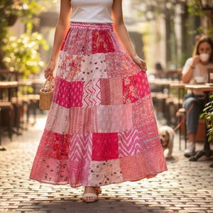 May include: A long, patchwork skirt in shades of pink, featuring diverse patterns and textures. The skirt is paired with a white top and sandals. A small woven basket is held. The scene is set on a cobbled street with outdoor seating, reminiscent of a cafe.