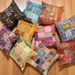 May include: A selection of square patchwork cushions in various colours, including blue, orange, yellow, and pink. The cushions display intricate patterns and textures. They are arranged on a wooden surface.