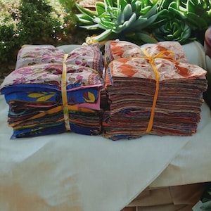 May include: Piles of patterned fabric squares, secured with yellow twine. The fabrics feature diverse patterns and colours, including floral and geometric designs. The stacks are arranged on a white surface, with greenery in the background.