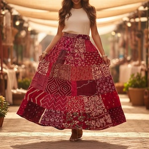 May include: A long, patchwork skirt in shades of red, burgundy, and pink. The skirt features various patterned fabric squares. The person is wearing a white top and brown sandals. The scene is set in a marketplace.