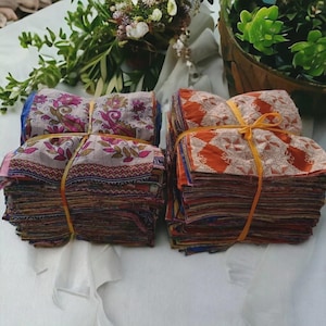 May include: Two stacks of patterned fabric squares, secured with yellow twine. One stack has floral designs in pink, purple, and beige. The other features an orange geometric pattern. The fabric is arranged on a white surface, with greenery in the background.