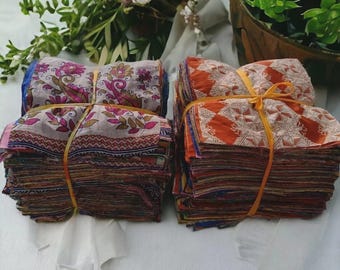 Könnte beinhalten: Zwei Stapel bunter Stoffquadrate, gebunden mit gelbem Garn. Ein Stapel zeigt florale Muster in Rosa, Lila und Beige. Der andere Stapel zeigt ein orangefarbenes geometrisches Design. Der Stoff liegt auf einer weißen Oberfläche, mit Grün im Hintergrund.
