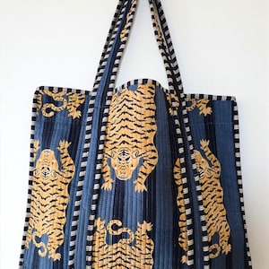 Op de afbeelding: Een gewatteerde draagtas met een marineblauw en wit gestreept patroon en tijgerillustraties in goud en zwart. De tas heeft bijpassende marineblauwe bandjes en een rechthoekige vorm.