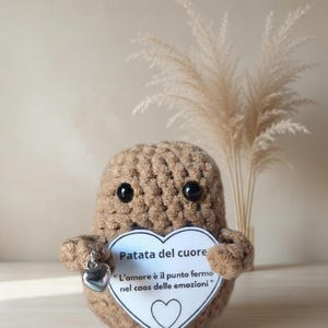 Può includere: Un peluche a forma di patata marrone all'uncinetto, con occhi a bottone neri. Tiene in mano un cartello bianco a forma di cuore con testo in italiano e un ciondolo a forma di cuore argentato. Erba della pampa decorativa sullo sfondo.
