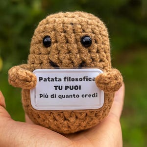 Puede incluir: Una figura de patata hecha a mano con ganchillo, con ojos de botón negros y una pequeña sonrisa. La patata sostiene un cartel que dice "Patata filosofica TU PUOI Più di quanto credi."