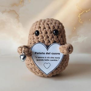 Puede incluir: Un peluche en forma de patata, tejido a ganchillo, en tonos marrones, con ojos de botón negros. Sostiene una tarjeta en forma de corazón con texto en italiano y un pequeño dije de corazón plateado. La tarjeta dice "Patata del cuore".