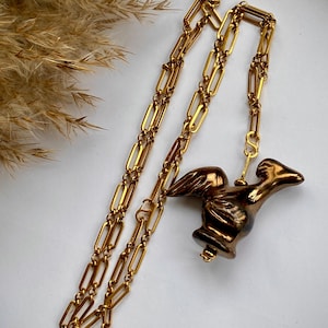 May include: A gold-toned necklace with a pendant shaped like a bird. The necklace features a long chain with rectangular links. The bird pendant is brown and gold, with detailed wings and a curved beak. The necklace is displayed on a white surface.