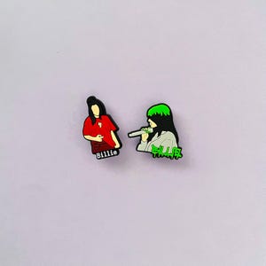 May include: Two colorful shoe charms featuring a singer. One charm depicts the singer in a red shirt with the name "Billie". The other charm shows the singer with green hair and the word "Killer".