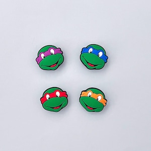 May include: Four colorful Teenage Mutant Ninja Turtles face charms. Each charm features a green turtle head with a different colored mask: purple, blue, red, and orange. The charms have a smiling expression and are set against a white background.