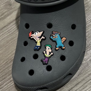 May include: A gray Croc shoe adorned with three colorful cartoon character charms. The charms feature characters in various poses, including a character with orange hair and a character wearing a hat.