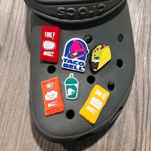 May include: A gray Crocs shoe adorned with colorful Taco Bell-themed charms. The charms include a red packet labeled "FIRE!", a Taco Bell logo, a taco, a green drink, an orange packet labeled "HOT", and a yellow packet labeled "(MILD)".