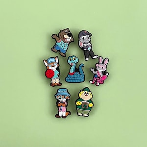May include: A collection of colorful cartoon character shoe charms. The charms feature various animals, including a beaver, rabbit, fox, snake, and bear, each wearing outfits and accessories. The charms are set against a solid green background.