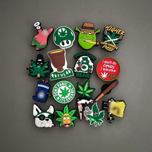 May include: A collection of colorful shoe charms with various designs, including cartoon characters, marijuana leaves, and humorous phrases like "Higher Than Giraffe Pussy" and "I don't do drugs I smoke weed."