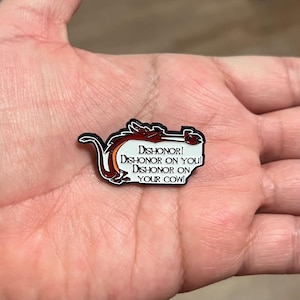 May include: A black, red, and white enamel pin featuring a dragon design with the text "Dishonor! Dishonor on you! Dishonor on your cow!" The pin has a black border and is likely a collectible item.
