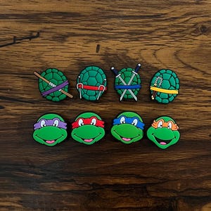 May include: A collection of eight Teenage Mutant Ninja Turtles-themed charms. The charms feature the turtles' faces with colored masks and their shells with weapons. The colors include green, purple, red, blue, and orange.