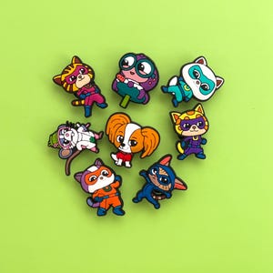 May include: A collection of colorful cartoon character charms, including cats, a dog, and a creature, arranged on a green background. Each charm is a different color and design, with superhero-themed costumes and masks.