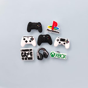 May include: A collection of gaming-themed charms, including black and white game controllers, a PlayStation logo, an Xbox logo, a pair of headphones, and a charm with the text "I PAUSED MY GAME TO BE HERE."
