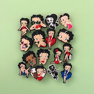 May include: A collection of colorful Betty Boop character charms in various poses and outfits. The charms feature black hair, red, pink, and blue dresses, and accessories. The background is a solid light green.