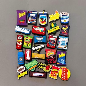 May include: A collection of colorful, miniature food-themed charms. The charms include snack bags, drink containers, and candy bars, with various brand logos and designs. The items are arranged on a gray surface.