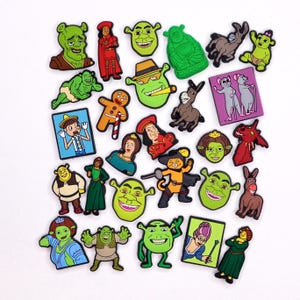 May include: A collection of colorful cartoon character charms, including Shrek, Donkey, and other characters. The charms are in various shapes and sizes, with vibrant green, brown, and red hues. The charms are likely for shoes or accessories.
