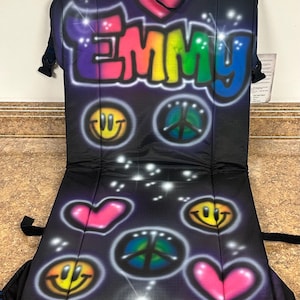 May include: A black folding chair with a colorful design featuring the name "EMMY" in rainbow letters, peace signs, and smiley faces. The chair has black straps.