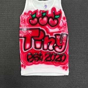 May include: White tank top with a red and black airbrushed design featuring three cherries and the text "Triz est 2020".