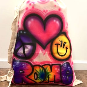 May include: A white canvas drawstring bag with a colorful spray-painted design. The design features a pink heart, a purple peace sign, a yellow smiley face, and the word "ROME" in rainbow colors.