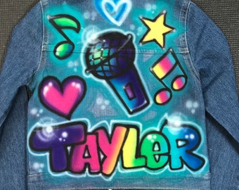 May include: A denim jacket with a vibrant airbrushed design. The back features a microphone, musical notes, hearts, a star, and the name "TAYLOR" in colorful graffiti-style lettering. The jacket has a classic collar and button closure.