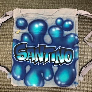 May include: A gray drawstring bag with a blue and teal paint splatter design. The name "Santino" is written in black and white graffiti style lettering.