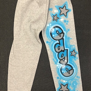 May include: Gray sweatpants with an elastic waistband and cuffs. The right leg features the word "dada" in a blue and gray graffiti-style design, surrounded by blue accents and gray stars.