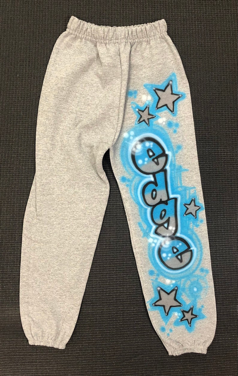 May include: Gray sweatpants with an elastic waistband and cuffs. The right leg features the word "dada" in a blue and gray graffiti-style design, surrounded by blue accents and gray stars.