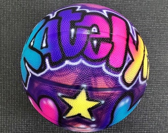 May include: A basketball with a vibrant graffiti design. The word "SAGE" is written across the ball in blue, purple, pink, and yellow. A yellow star is featured. The ball has a textured surface.