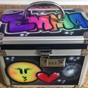 May include: A silver metal case with a combination lock. The case has a colorful graffiti design with the name "EMMA" on the top and a smiley face and a red heart on the bottom.