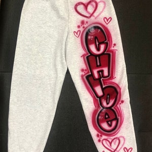 May include: Light gray sweatpants with the name "Chloe" in a graffiti-style design in red and black. The design includes hearts and is airbrushed onto the left leg. The sweatpants are made of a soft material.