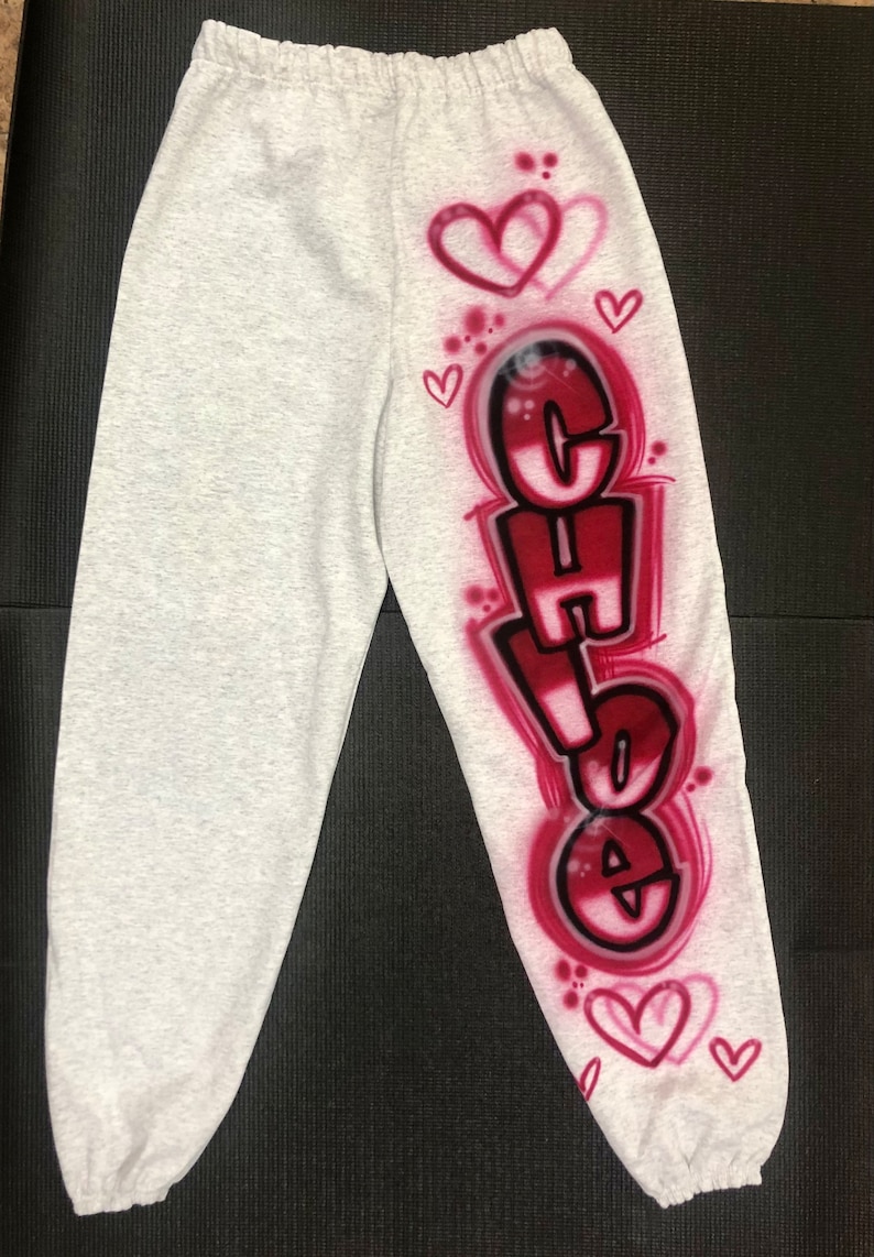 May include: Light gray sweatpants with the name "Chloe" in a graffiti-style design in red and black. The design includes hearts and is airbrushed onto the left leg. The sweatpants are made of a soft material.
