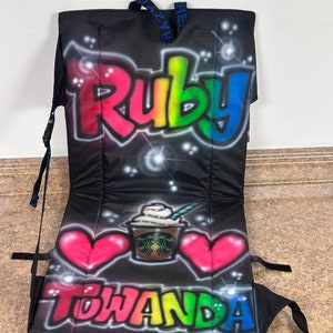 May include: A black folding chair with a personalized design. The chair features a rainbow-colored graffiti-style name "Ruby" at the top, a Starbucks coffee cup with whipped cream and a straw in the center, and the name "Towanda" in rainbow-colored graffiti-style lettering at the bottom. Two pink hearts are on either side of the coffee cup.