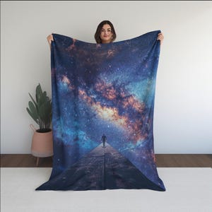 May include: A large, rectangular blanket featuring a vibrant depiction of a starry galaxy. The design includes a figure walking on a path towards the cosmos, with shades of blue, purple, and orange. The blanket is held up by a person.