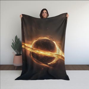 May include: A black blanket featuring a vibrant depiction of a black hole with a bright orange and yellow ring of light. The blanket has a dark background with a celestial theme.