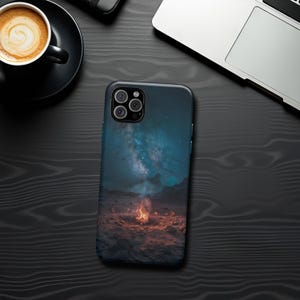 May include: A smartphone case featuring a vibrant image of a campfire under a starry night sky. The case has a dark blue and orange color scheme, with a detailed depiction of the Milky Way. The case is on a dark wooden surface, next to a cup of coffee and a laptop.