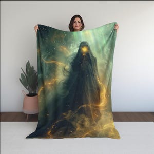 May include: A dark blanket featuring a hooded figure with a glowing face, set against a backdrop of swirling green and gold nebulas. The design evokes a sense of mystery and fantasy, with a celestial theme.