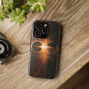 Puede incluir: Funda de teléfono inteligente negra con un diseño celestial. Presenta dos círculos oscuros y una brillante explosión de luz naranja y amarilla en el centro. La funda está sobre una superficie de madera.