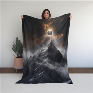 Könnte beinhalten: Eine rechteckige Wohndecke mit einem kontrastreichen Grafikdruck eines schneebedeckten Berggipfels unter einem dunklen Himmel. Die Szene zeigt eine Sonnenfinsternis mit einem leuchtenden Sonnenring, umgeben von orangefarbenen Wolken und Nebel.